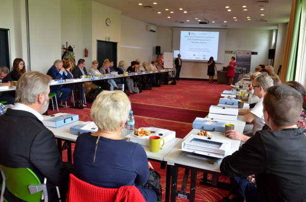 Początek debaty. Widok z rogu sali konferencyjnej. Uczestnicy usadzeni w podkowie. Prowadzący stoją przodem do uczestników.