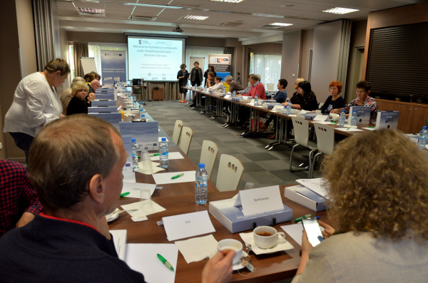 Widok na salę konferencyjną z lewego rogu sali. Uczestnicy usadzeni w podkowie. Początek debaty. Prowadzący stoją przed ekranem przodem do uczestników.